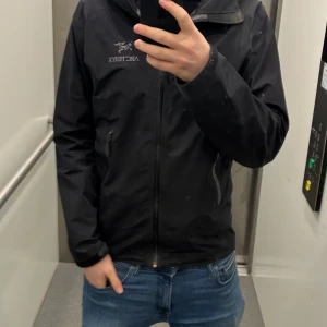 Svart vindjacka från Arc'teryx - Säljer en svart vindjacka från Arc'teryx med dragkedja framtill och två sidofickor. Jackan har hög krage och lång ärm, perfekt för blåsiga dagar. Enkel och stilren design med Arc'teryx-logga på bröstet.