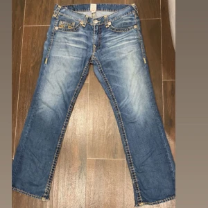 True Religion - Säljer ett par klassiska blå jeans från True Religion med snygga slitningar och kontrastsömmar. Byxorna har raka ben men är baggy