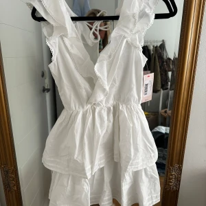 Studentklänning  från Nelly - Supersöt vit klänning från Nelly med volangdetaljer och djup v-ringning. Klänningen har ärmlös design, knytning i ryggen och dubbla lager i kjolen för extra volym. Perfekt för sommarens alla tillfällen!