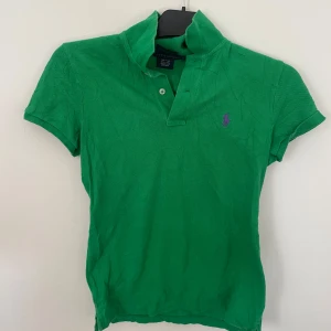 Grön pikétröja från Ralph Lauren - Säljer en klassisk grön pikétröja från Ralph Lauren i 100% bomull. Tröjan har korta ärmar, krage och två knappar framtill. Liten broderad logga i lila på bröstet. Perfekt för en sportig och stilren look!