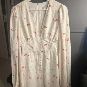 Vit blommig långärmad klänning från Adoore - Superfin vit kort klänning från Adoore med små röda blommor, v-ringning och knappar framtill. Långärmad modell i strukturerat tyg som ger en lyxig känsla. Den är i väldigt bra skick och perfekt för vår och sommar!