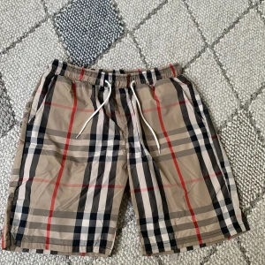Beige burberry shorts  - Snygga beige burberry shorts med klassiskt rutig mönster i svart, vitt och rött. De har elastisk midja med vit snörning och är knälånga. Perfekta för sommaren och har en avslappnad passform.