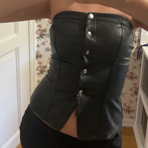 Svart skinnkorsett med knappar - Säljer en helt ny oanvänd svart axelbandslös korsett i skinnimitation med knappar framtill. Snygg och tight passform som framhäver midjan. Från H&M i storlek Xs men passar även S. Aldrig använd då jag är lite obekväm i den. Skönt material annars!