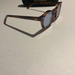 solglasögon från TBD Eyewear - Snygga runda solglasögon med brunskiftande bågar och blåtonade linser. Bågarna har en klassisk, tidlös design i acetat och passar perfekt för dig som gillar en stilren look. Märket TBD Eyewear syns på fodralet.