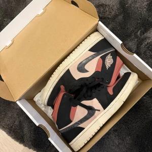 Nike Air Jordan 1 Mid i svart, rosa och beige - Säljer ett par Nike Air Jordan 1 Mid i snygg färgkombination av svart, rosa och beige. Skorna har klassisk snörning, hög modell och ikoniska Air Jordan-loggan på sidan. Perfekta för dig som vill sticka ut med dina sneakers.