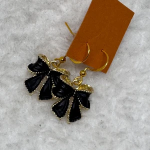 Svarta rosettörhängen med gulddetaljer - Supersnygga örhängen med svarta rosetter och guldfärgade detaljer. Hängande modell som ger en cool och trendig look. Perfekta accessoarer för att lyfta din outfit med en unik touch. Krokarna är i sterling silver 