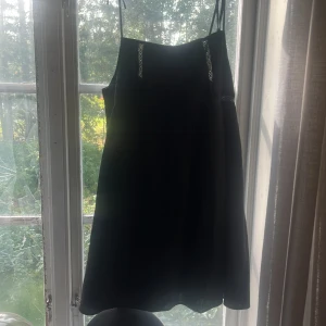 Svart kort klänning med spetsdetaljer - Säljer en svart kort klänning med tunna axelband som knyts upptill. Klänningen har fina spetsdetaljer framtill och ett subtilt broderat mönster på bröstet. Passar dig som gillar stilrena plagg med lite extra detaljer. Köpt för 250, aldrig använd 