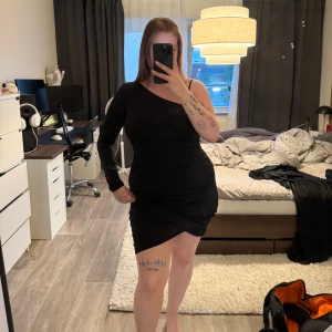 Svart oneshoulder klänning NA-KD XL -  Klänningen har en unik oneshoulder-design med en lång ärm och öppen på andra sidan. Figurnära passform och stretchigt material som känns bekvämt.