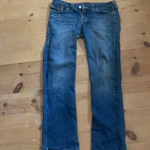 True Religion blå jeans, stl 31 - Säljer ett par klassiska blå jeans från True Religion med snygga kontrastsömmar och ikoniska bakfickor med lock och silverfärgade knappar. Jeansen har rak passform och normal midja. Tillverkade i USA och i mjukt denimtyg.