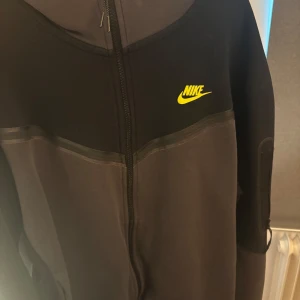 Svart och grå hoodie från Nike - Nike hoodie i svart och mörkgrått med dragkedja och gul logga på bröstet. Tröjan har huva och är långärmad, perfekt för chill dagar eller när du vill ha en sportig look. Materialet är mjukt och skönt, troligtvis bomull och polyester.