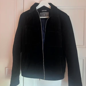 Svart manchesterjacka Human Scales - Snygg svart mocka jacka från Human Scales, modell Rick. Jackan har klassisk krage, två bröstfickor och dragkedja framtill. Perfekt för dig som gillar stilren och tidlös look med lite edge. Nypris 4999kr mitt pris 699kr 