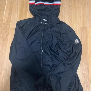 Svart vindjacka från Moncler med huva - Svart vindjacka från Moncler med klassisk logga på ärmen. Jackan har huva med snygga röd, vit och blå ränder, dragkedja framtill och snörning vid halsen. Tillverkad i lätt och glansig polyester, perfekt för dig som vill ha en stilren och sportig look.