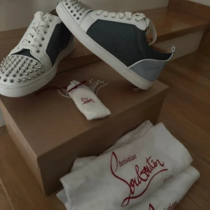 Christian Louboutin sneakers med nitar - Snygga sneakers från Christian Louboutin i vitt och grått med silverfärgade nitar på tån, röda sulor och detaljer i skinn och mocka. Storlek 43,5 men passar mer som 44. Skorna har snörning och en cool, edgy vibe som sticker ut. Perfekta för dig som vill ha något unikt och lyxigt i garderoben. 10/10 i skick, Knappast använda och kvitto medföljer. Tar gärna byten också sålänge de är rimliga