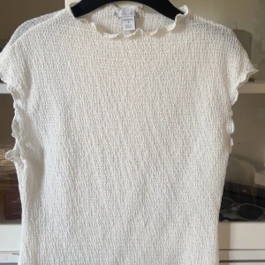 Vit strukturerad topp från New yorker  - Superfin vit topp från New yorker med strukturerat tyg och små volangdetaljer vid halsringningen. Ärmlös modell med lätt croppad känsla, perfekt till jeans eller kjol. Enkel men ändå söt och passar till många olika stilar.