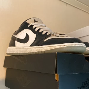 Nike Air Jordan 1 Low svart/vit - Snygga Nike Air Jordan 1 Low sneakers i svart och vitt med klassisk swoosh på sidan. Skorna har snörning, rund tå och platt sula. Perfekta för dig som gillar streetwear och vill ha en ikonisk look.