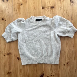 Beige stickad tröja med puffärm Vero Moda - Superfin beige stickad tröja från Vero Moda med korta puffärmar och ribbade muddar. Perfekt för dig som vill ha en söt och trendig look med lite volym på axlarna. Enkel att matcha med både jeans och kjol.