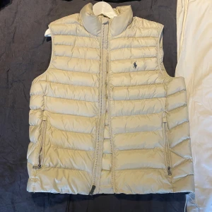 Beige dunväst från Polo Ralph Lauren - Snygg beige dunväst från Polo Ralph Lauren med hög krage och två dragkedjefickor framtill. Västen har quiltad design och klassisk logga broderad på bröstet. Perfekt att slänga över en hoodie eller tröja för extra värme. Inte använd. Ny pris 5100kr.