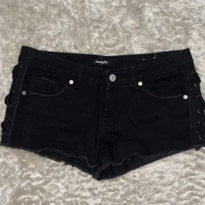 Svarta jeansshorts med snörning  - Svarta jeansshorts från Jennyfer med snygg snörning på sidorna och rå kant nedtill. Klassisk femficksmodell, silverfärgade nitar och knapp. Låg/ medel midja. Det är storlek 34 men skulle säga att den kan passa S! Midjemått: 39cm