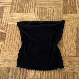 Svart tubtopp från Gina Tricot M - En svart tubtopp från Gina Tricot i storlek M. Toppen är axelbandslös med en enkel och stilren design, har lätt vågig kant nertill och är gjord i ett stretchigt material som sitter snyggt på kroppen. Perfekt för att skapa en clean och trendig look.