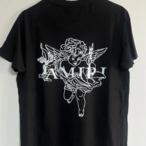 Svart Amiri t-shirt med ängeltryck - Svart t-shirt från Amiri i mjuk bomull med stort vitt ängeltryck och logga på ryggen samt färgstänk och logga framtill. Klassisk rund hals och korta ärmar. Perfekt för dig som vill sticka ut med en cool streetstyle-look.