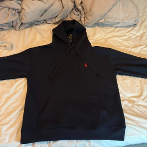 Mörkblå hoodie från Polo Ralph Lauren - Säljer en mörkblå hoodie från Polo Ralph Lauren med klassisk röd logga broderad på bröstet. Tröjan har huva med snörning, magficka och ribbade muddar. Perfekt för dig som gillar stilren och enkel design med premiumkänsla.