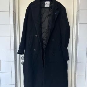 Svart lång oversized kappa från Zara - Säljer en svart lång oversized kappa från Zara med klassisk krage och dubbelknäppning. Kappan har raka linjer, två fickor framtill och är helfodrad. Perfekt för dig som gillar en stilren och tidlös look under höst och vinter. Blåa detaljer, ett litet hål på ryggen, syns knappt