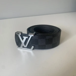 Louis Vuitton svart rutigt bälte - Snyggt svart bälte från Louis Vuitton med klassiskt rutigt mönster och stor LV-logga i silvermetall framtill. Bältet är gjort i skinn och har exklusiva detaljer med Louis Vuitton-gravyr på spännet. Perfekt accessoar för att lyfta din outfit.