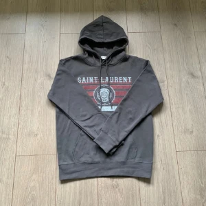 Grå Saint Laurent hoodie med tryck - Säljer en grå hoodie från Saint Laurent med coolt tryck i vitt och rött på bröstet. I storlek L. Tröjan är i Nyskick och jag har kvitto, skriv om du har några frågor 