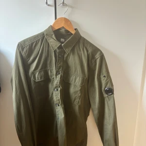 Olivgrön skjorta från C.P. Company - Säljer en olivgrön skjorta från C.P. Company med vanlig krage, bröstfickor och knappar framtill. Skjortan har lång ärm och badge på vänster ärm. 