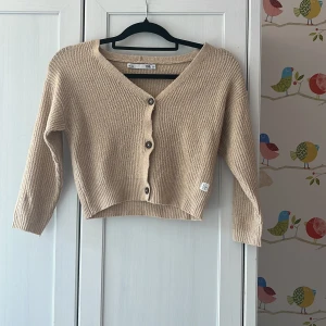 Beige stickad kofta från 157 - Superfin beige stickad kofta från 157 med v-ringning och tre knappar framtill. Modellen är croppad och har långa ärmar. Perfekt att slänga över en t-shirt eller linne för en chill look. Liten patch med texten 'ETT FEM SJU', inte jätte använd