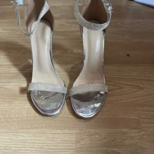 Glittriga silverfärgade klackar - Snygga klackskor i silver med transparent blockklack och glittrande remmar över tårna och runt ankeln. Skorna har öppen tå och spänne vid ankeln. Perfekta för att sticka ut och ge extra bling till outfiten.