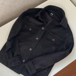 Svart mocka overshirt från Zara - Säljer en svart overshirt från Zara i mjuk mocka-look. Jackan har klassisk krage, knäppning framtill med knappar och två stora bröstfickor med lock. Perfekt att slänga över en hoodie eller t-shirt för en clean och stilren vibe.