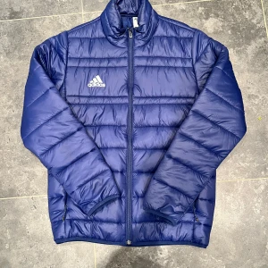 Adidas jacka  - Snygg blå pufferjacka från Adidas med vitt logotryck på bröstet. Jackan har dragkedja framtill, ståkrage och två sidofickor med dragkedja. Perfekt för kyliga dagar och har en clean, sportig look som funkar till det mesta.