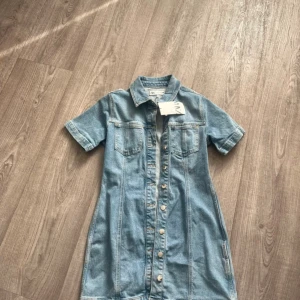 Jeansklänning med knappar från Zara - Snygg jeansklänning från Zara i ljusblå denim med kort ärm, krage och knappar hela vägen framtill. Två bröstfickor ger en cool touch och materialet är klassiskt jeans. Perfekt för dig som gillar en avslappnad men trendig stil.