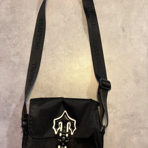 Svart axelväska från Trapstar - Säljer en svart axelväska från Trapstar med justerbart band där loggan är invävd. Väskan har lock med unik vit och svart detalj framtill, spänne och dragkedja. Materialet är slitstark polyester med diskret mönstrad framsida. Perfekt för att bära det viktigaste.