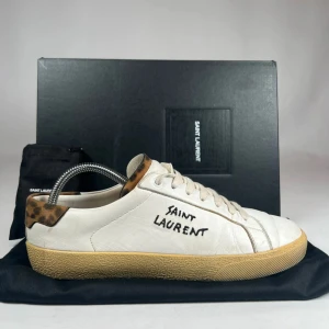 Saint Laurent vita sneakers med leoparddetalj - Saint Laurent sneakers i vitt skinn med handskriven logga på sidan och cool leopardmönstrad detalj vid hälen. Klassisk rund tå, beige sula och snörning framtill. Perfekt för dig som vill ha en unik och trendig look.