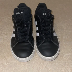 Svarta Adidas sneakers med vita ränder - Snygga svarta Adidas sneakers med klassiska vita ränder på sidorna och vit sula. Skorna har snörning och är tillverkade i syntetmaterial med en rund tå. Perfekta för dig som gillar en clean och sportig stil.