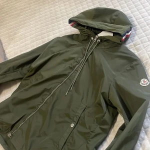Grön vindjacka från Moncler med huva - Snygg mörkgrön vindjacka från Moncler med huva och dragsko. Jackan har dragkedja framtill, Moncler-logga på ärmen och coola ränder på insidan av huvan. Perfekt för dig som vill ha en stilren och sportig look. Stl 3 som motsvarar ungefär L! 