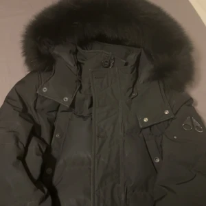 Svart dunjacka med päls från Moose Knuckles - Säljer en svart dunjacka från Moose Knuckles med stor fluffig päls runt huvan och ikonisk logga på ärmen. Jackan har dragkedja, tryckknappar och flera fickor. Perfekt för kalla vinterdagar och riktigt snygg streetstil.