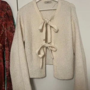 Krämvit stickad kofta med knyt - Supermysig krämvit stickad kofta från Twisted Gallery med två stora knytband framtill. Koftan har ribbad struktur och långa ärmar, perfekt för lager-på-lager och chill dagar. Snygg att matcha med jeans eller kjol för en soft look.