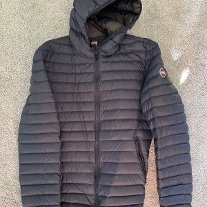 Mörkblå Colmar pufferjacka med huva - Säljer en mörkblå pufferjacka från Colmar med huva och dragkedja framtill. Jackan har horisontella quiltade sömmar, två fickor med dragkedja och en rund Colmar-logga på ärmen. Perfekt för kyliga dagar och riktigt snygg streetstil. Nyskick, använd ett fåtal gånger! Storlek 50