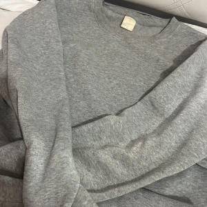 Grå sweatshirt i mjuk bomull - En basic grå sweatshirt med rund hals och långa ärmar. Tröjan är i mjuk bomull och har en klassisk passform som funkar till allt. Perfekt för chill dagar eller när du vill ha något enkelt och bekvämt.