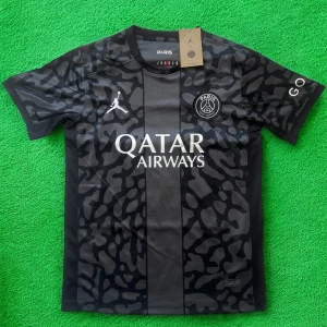 PSG x Jordan svart fotbollströja - Snygg svart PSG fotbollströja i samarbete med Jordan. Tröjan har korta ärmar, coolt kamouflagemönster och stora loggor från både PSG och Jordan på bröstet. Tillverkad i lätt och ventilerande material, perfekt för match eller träning.