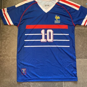 Frankrike Zidane VM 98 fotbollströja - Säljer en ikonisk blå Frankrike fotbollströja från VM-finalen 1998 med nummer 10 och Zidane på ryggen. Tröjan har vita och röda detaljer på ärmarna, broderat tupp-emblem med datumet för finalen och klassiska ränder framtill. Perfekt för dig som älskar retro och fotbollshistoria.