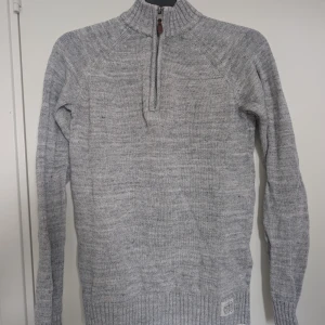 Grå stickad tröja med half zip - Snygg grå stickad tröja från Peak Performance med ribbad krage och half zip-dragkedja. Långärmad modell med diskret logga nertill. Strl 170 motsvarar en S i herr. 