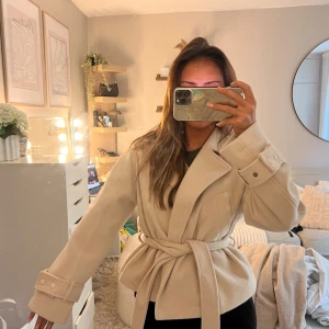 Beige kappa med bälte från Nelly - Snygg beige kappa från Nelly med bred krage och knytskärp i midjan. Jackan har långa ärmar med dekorativa spännen vid ärmsluten och en klassisk omlottstil🌸