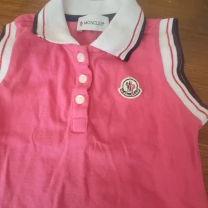 Rosa ärmlös polotopp för Barn, från Moncler storlek 92 - Rosa ärmlös polotopp för Barn, från Moncler med vit krage och knäppning framtill. Toppen är använd 2-3 ggr, så är i väldigt bra skick. Storlek 92.