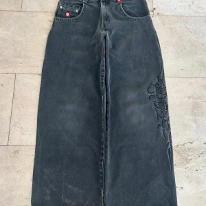Nyss skickade från USA Kom och hämta dina JNCO-jeans - Riktigt feta svarta JNCO jeans med bred passform och tribal brodyr längs benet och bakfickan. Klassisk femficksmodell i denim med coola detaljer och tydlig streetwear-vibe. Perfekta för dig som gillar baggy stil och vill sticka ut.