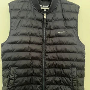 Mörkblå dunväst från Gant - Snygg svart dunväst från Gant med quiltad design och dragkedja framtill. Västen har två fickor på sidorna och diskret Gant-logga på bröstet. Perfekt att slänga över en hoodie eller tröja för en sportig och clean look.