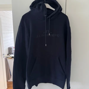 Blå hoodie från J.Lindeberg - Säljer en marinblå hoodie från J.Lindeberg med broderad logga på bröstet. Tröjan har huva med snörning och en stor magficka. Perfekt för dig som gillar en stilren och enkel look.